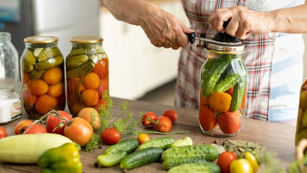 Eine Person verschließt mit einer Dosenverschlussmaschine ein Glas mit eingelegten Gurken und Tomaten.