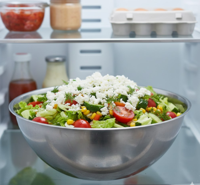 Ein fertig angerichteter, bunter Partysalat in einer großen Edelstahlschüssel auf einer hellen Küchenoberfläche. Der Salat ist reichhaltig mit Feta-Käse bestreut und wird gerade mit frischen, grünen Kräutern (wie Petersilie oder Basilikum) garniert. Im Hintergrund sieht man einen modernen Kühlschrank, was andeutet, dass der Salat nun zum Durchziehen kühl gestellt wird. Das Bild vermittelt Frische und eine appetitliche Optik.