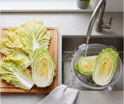 Draufsicht auf eine Küchenspüle, in der Chinakohl vorbereitet wird. Ein halbiertes Stück Kohl liegt in einer Salatschleuder unter fließendem Wasser, während auf einem Holzschneidebrett daneben weitere Blätter und die andere Kohlhälfte liegen.