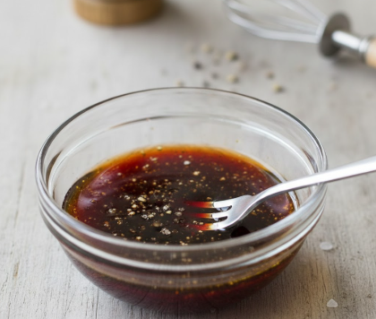 Eine kleine Glasschale mit einer dunklen Vinaigrette aus Olivenöl und Rotweinessig, gewürzt mit Meersalz und Pfeffer. In der Schale steckt eine Gabel zum Aufschlagen, im Hintergrund sind ein Schneebesen und Gewürze angedeutet.