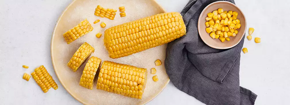 Frischer gelber Maiskolben, teilweise in Stücke geschnitten, auf einem Teller neben einer Schale mit Maiskörnern und einem grauen Küchentuch.