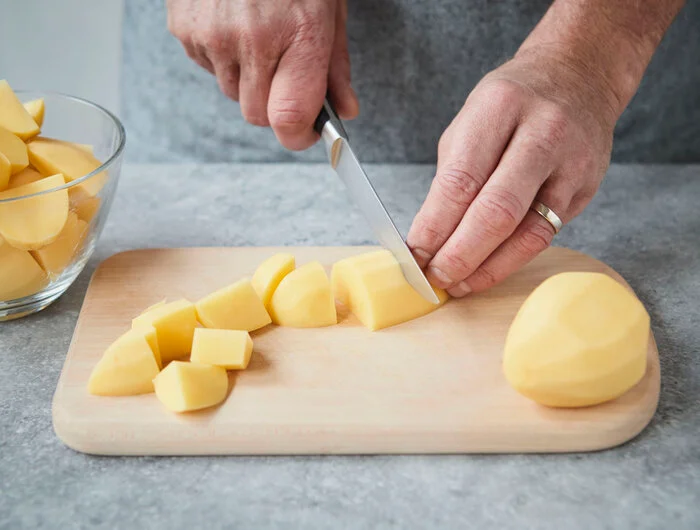 Eine Person schneidet geschälte Kartoffeln mit einem Messer auf einem Holzbrett in Würfel.