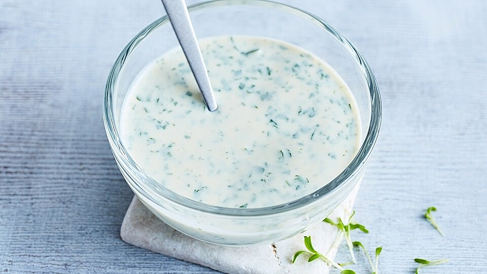 Eine Glas-Schüssel mit cremiger weißer Kräutersauce oder Joghurtdressing, in der ein silberner Löffel steckt, auf einem hellen Hintergrund.