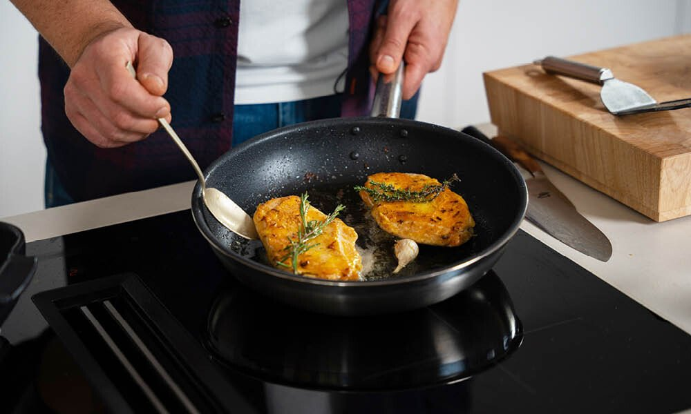 Eine Person brät zwei goldbraun marinierte Hähnchenbrustfilets in einer schwarzen Pfanne auf einem modernen Induktionsherd an. Die Filets sind mit frischen Kräutern (Rosmarin und Thymian) sowie einer Knoblauchzehe garniert. Mit einem Löffel wird das Fleisch während des Bratvorgangs mit Bratfett übergossen. Im Hintergrund sind ein massives Holzschneidebrett und Küchenmesser erkennbar.