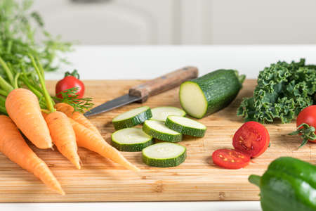 Ein helles Holzschneidebrett mit einer Auswahl an frischem Gemüse: Ganze Karotten mit Grün, in Scheiben geschnittene Zucchini, halbierte Kirschtomaten, Grünkohl und eine grüne Paprika liegen dekorativ um ein kleines Küchenmesser mit Holzgriff verteilt.