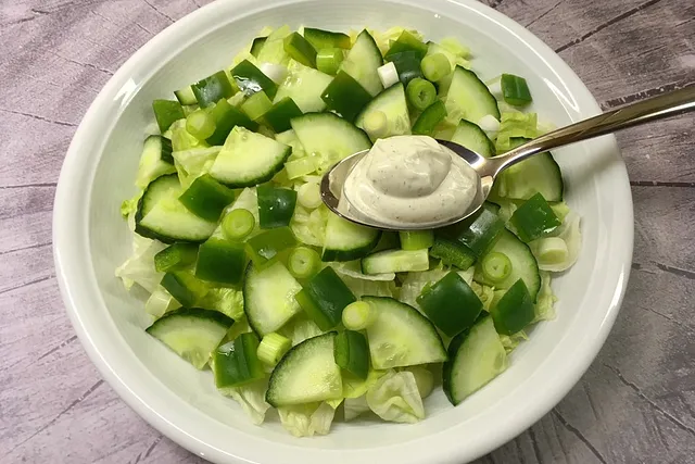 Eine weiße Schüssel mit einem frischen Salat aus hellem Blattsalat, gewürfelten grünen Paprikastücken, Gurkenscheiben und Frühlingszwiebeln. Ein silberner Löffel mit einem Klecks weißem cremigen Dressing liegt oben auf dem Gemüse.