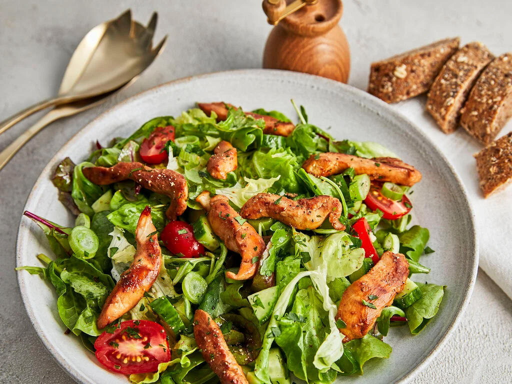 Ein appetitlich angerichteter bunter Salat in einer hellen Schale, getoppt mit gebratenen Hähnchenstreifen. Der Salat besteht aus grünem Blattsalat, halbierten Kirschtomaten, Gurkenscheiben und Frühlingszwiebeln. Im Hintergrund sind goldenes Besteck, eine hölzerne Pfeffermühle und Scheiben von dunklem Vollkornbrot zu sehen.