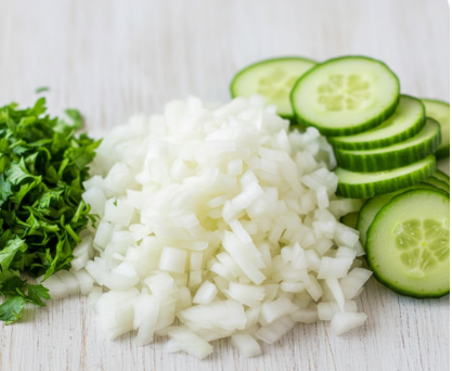 Frisch vorbereitete Kochzutaten auf einer hellen Holzoberfläche: Ein großer Haufen fein gewürfelter weißer Zwiebeln in der Mitte, daneben ordentlich aufgestapelte grüne Gurkenscheiben auf der rechten Seite und ein Bett aus fein gehackter frischer Petersilie auf der linken Seite.