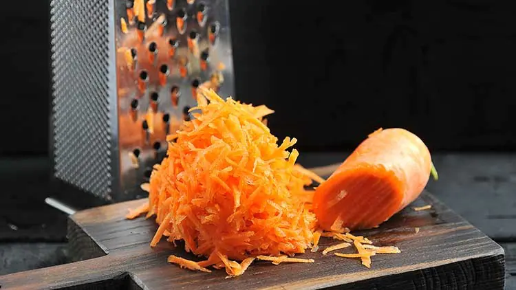 Frisch geriebene Karotten auf einem dunklen Holzbrett neben einer Küchenreibe.