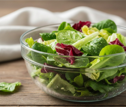 Ein frischer, bunter Blattsalat in einer Glasschüssel, die auf einem rustikalen Holztisch steht. Die Salatblätter sind mit Wassertropfen benetzt und bestehen aus einer Mischung von grünem Feldsalat und rötlichem Radicchio. Im Hintergrund ist ein helles, weiches Tuch unscharf zu erkennen.